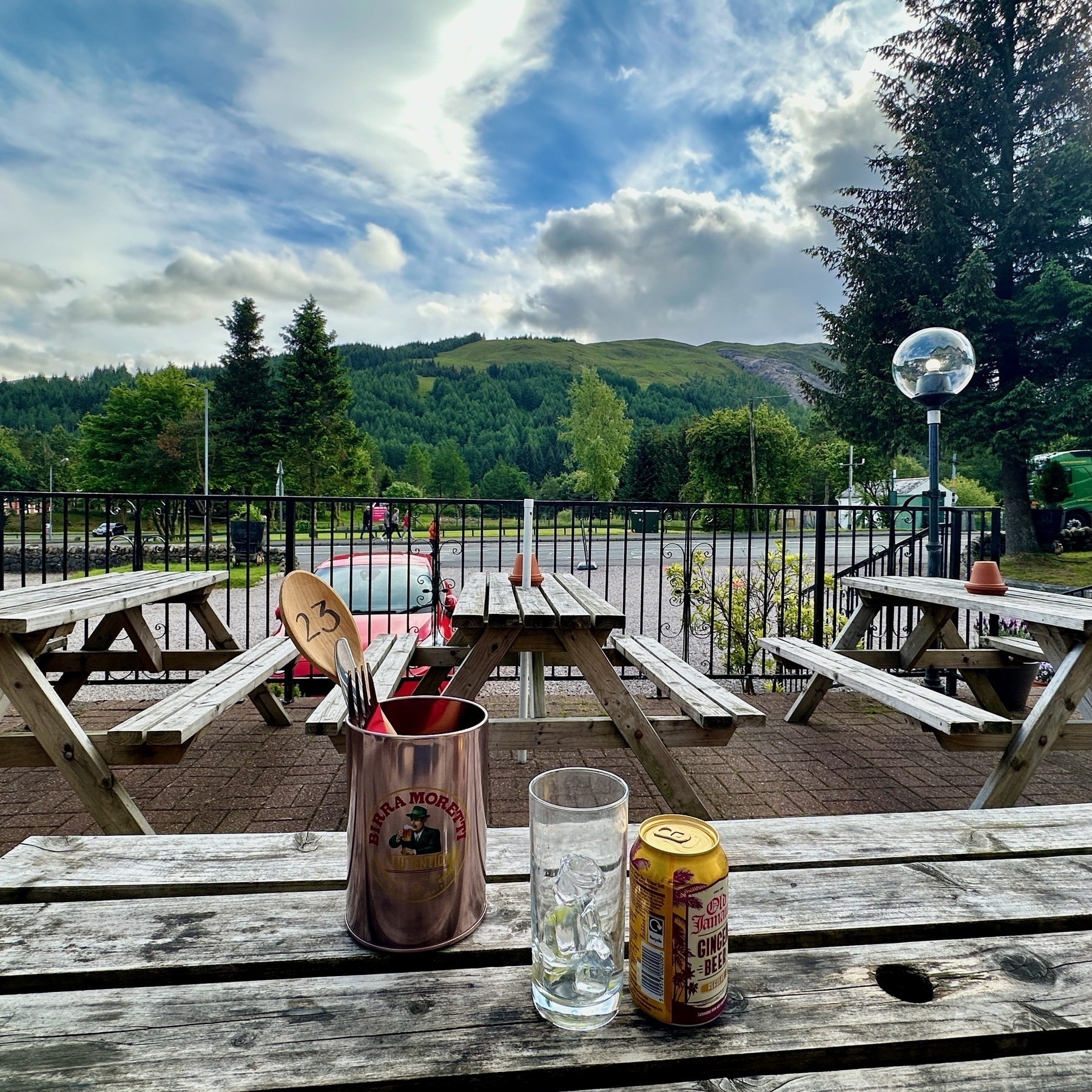 Auto-generated description: A rustic outdoor seating area features wooden picnic tables with a scenic view of lush green mountains under a partly cloudy sky.