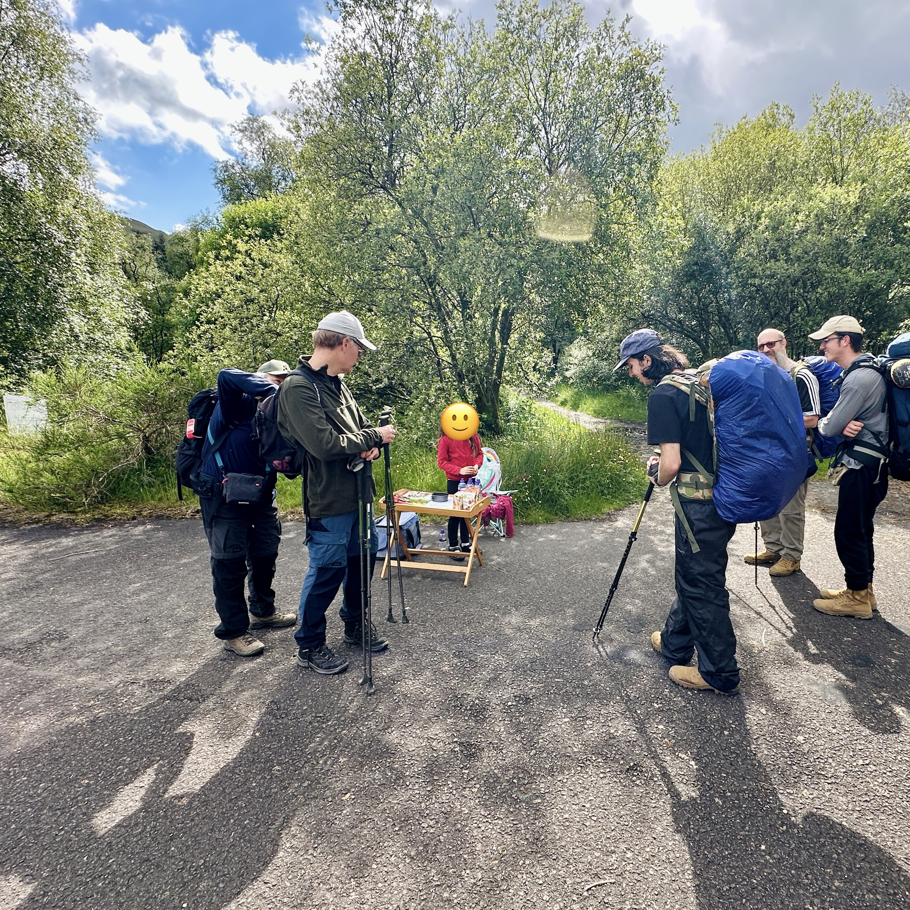 Auto-generated description: A group of hikers with backpacks and trekking poles are gathered around a small table with a chair, set up with snacks and beverages, near trees and bushes.