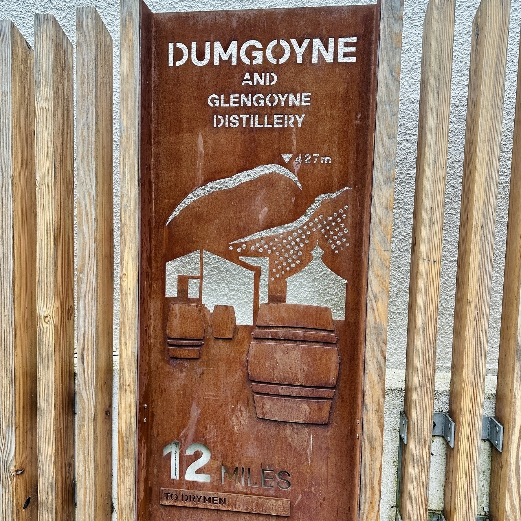 Auto-generated description: A wooden sign depicts Dumgoyne and the Glengoyne Distillery, indicating it is 12 miles to Drymen, with images of barrels, buildings, and a mountain backdrop.