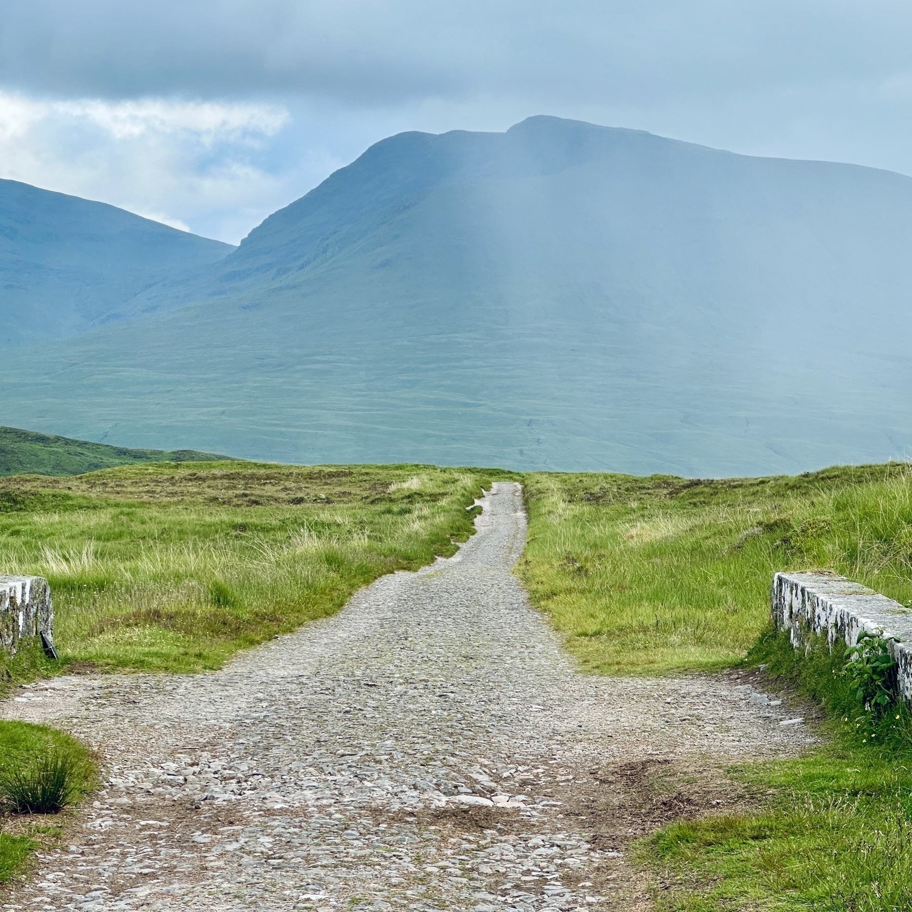 Auto-generated description: A narrow gravel path stretches into the distance between grassy fields, leading towards mist-covered mountains under a cloudy sky.