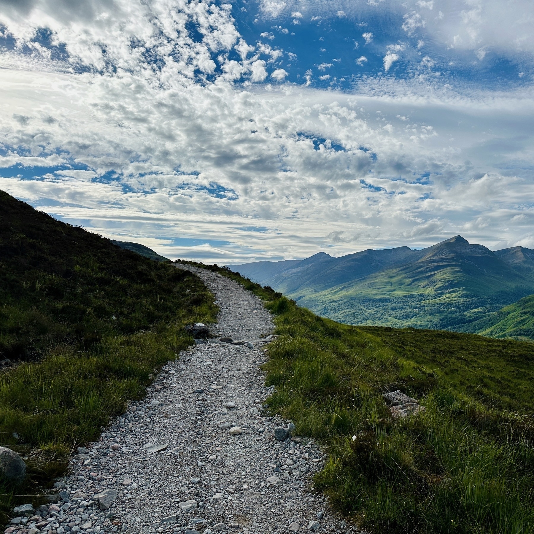 Auto-generated description: A rocky path winds through a lush, green hillside under a sky filled with dramatic, scattered clouds, leading towards distant mountains.