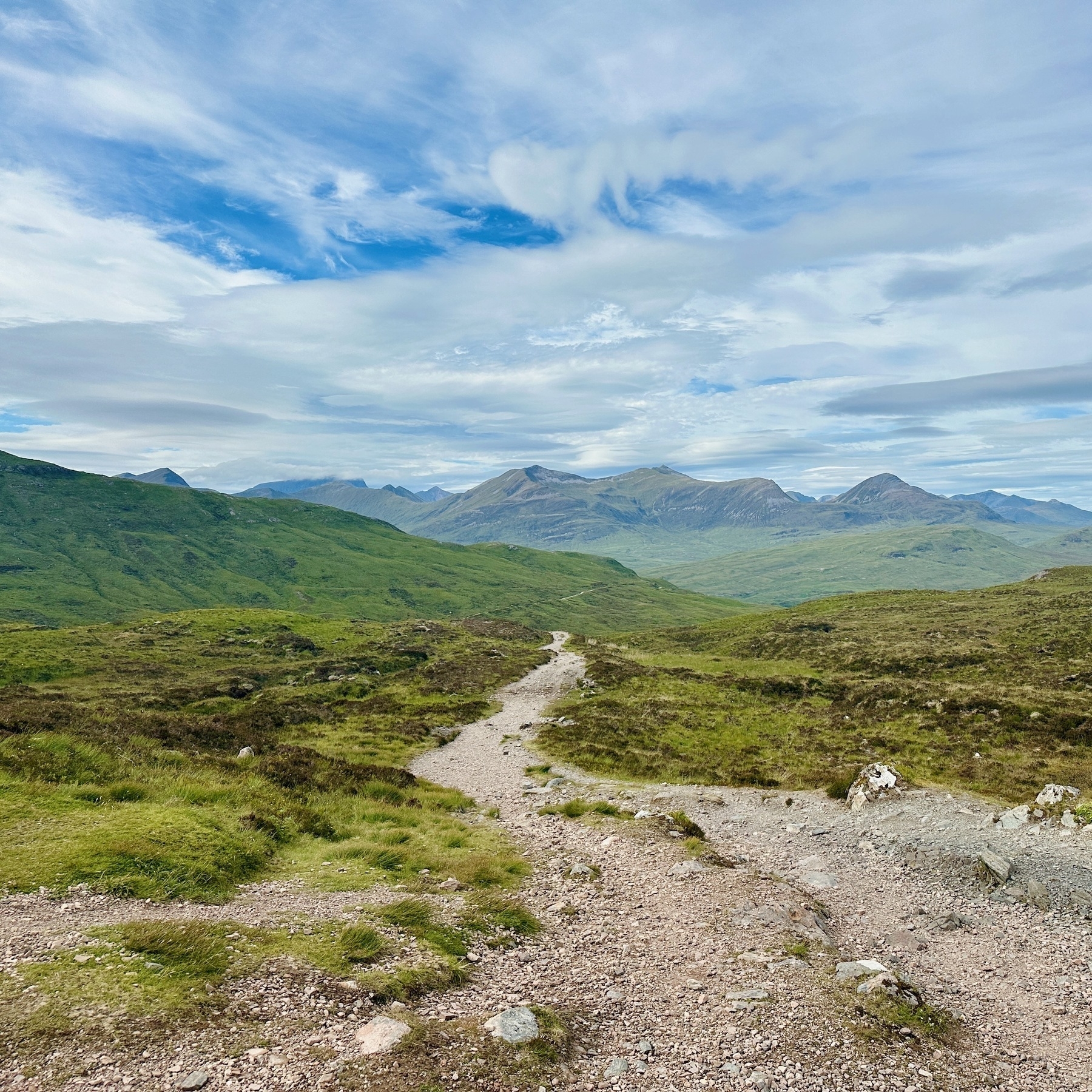 Auto-generated description: A winding dirt path cuts through a lush green landscape with rolling hills and distant mountains under a partly cloudy sky.