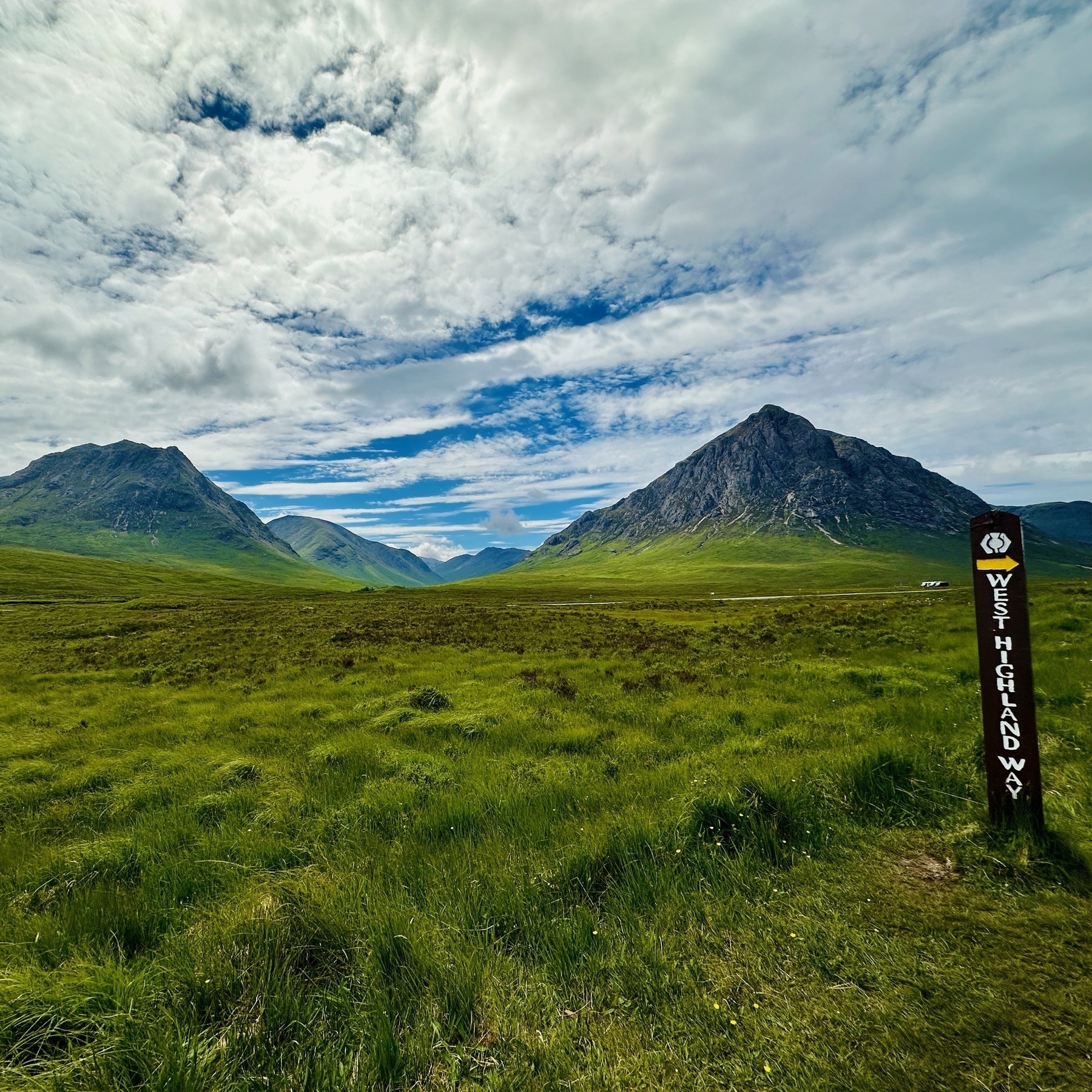 Auto-generated description: A lush, green, scenic landscape features the towering peaks of mountains under a partly cloudy sky, with a signpost on the right indicating the West Highland Way.