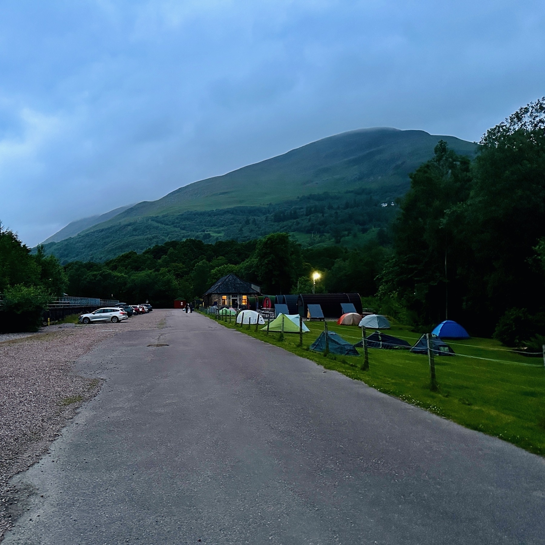 Auto-generated description: A serene campsite with several tents and a few cars is situated alongside a gravel path with a mountainous landscape in the background during dusk.