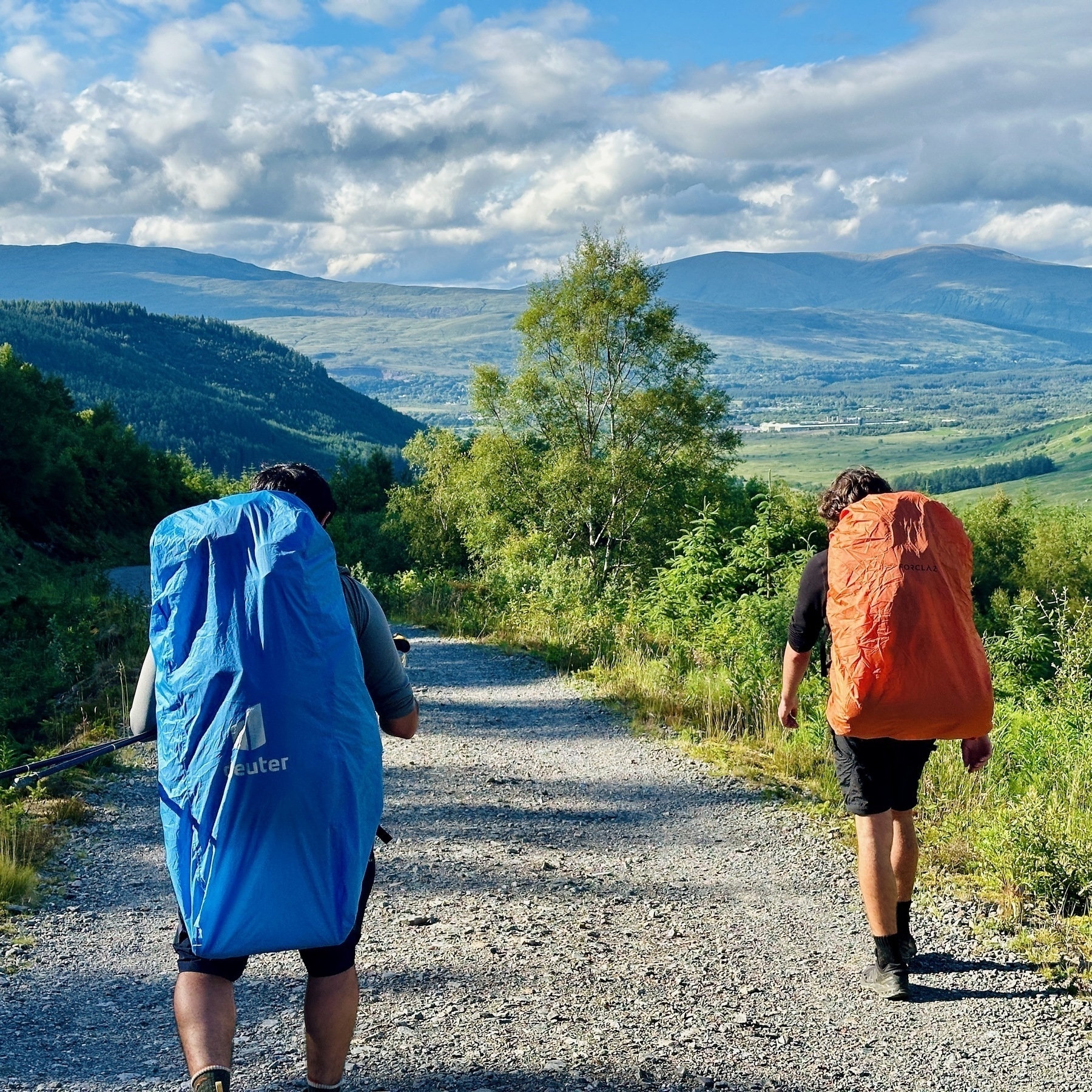 Auto-generated description: Two hikers with large backpacks are walking along a gravel path in a scenic, mountainous landscape with lush greenery and a partly cloudy sky.