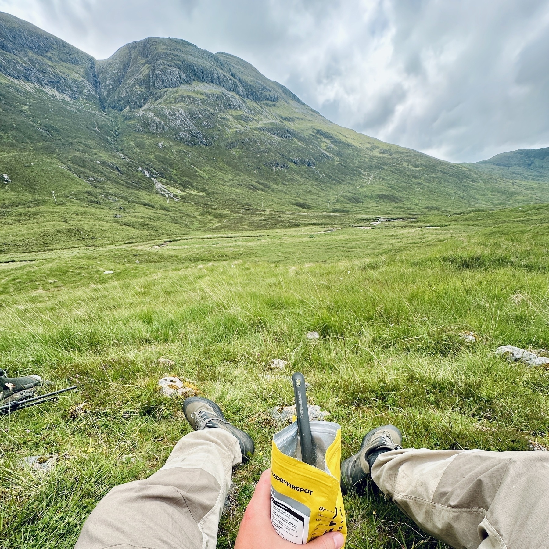 Auto-generated description: A person is sitting in a grassy field with mountains in the background, holding a yellow rehydrated meal package in their hand.