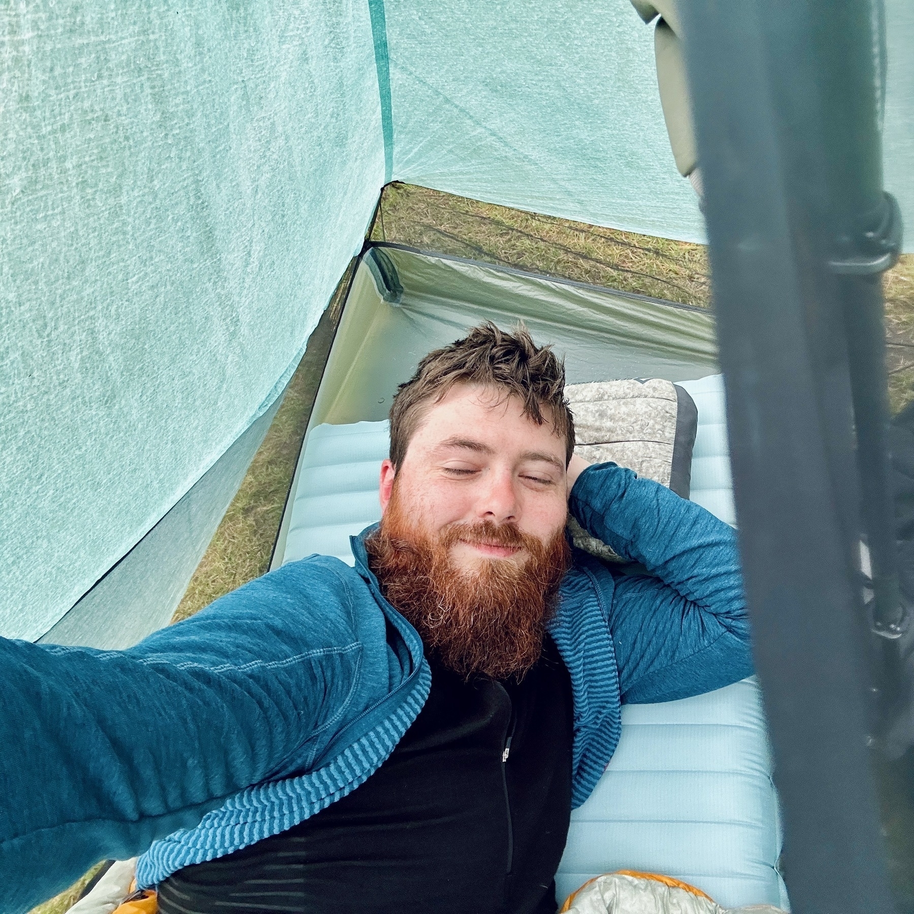 Auto-generated description: A bearded man wearing a blue jacket is lying down and smiling inside a tent, possibly taking a selfie.