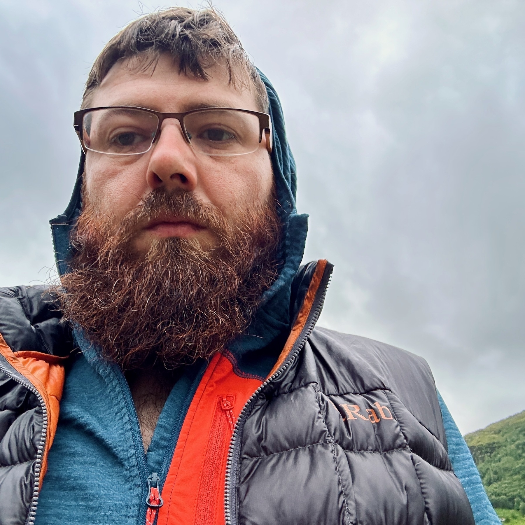 Auto-generated description: A bearded man with glasses is wearing a hooded jacket and looking directly at the camera against an outdoor background.