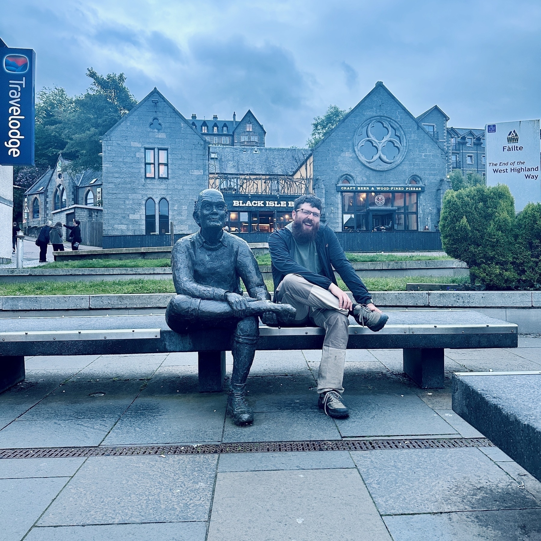 Me sitting on the bench that marks the new end of the West Highland Way, with the statue of the man rubbing his sore feet.