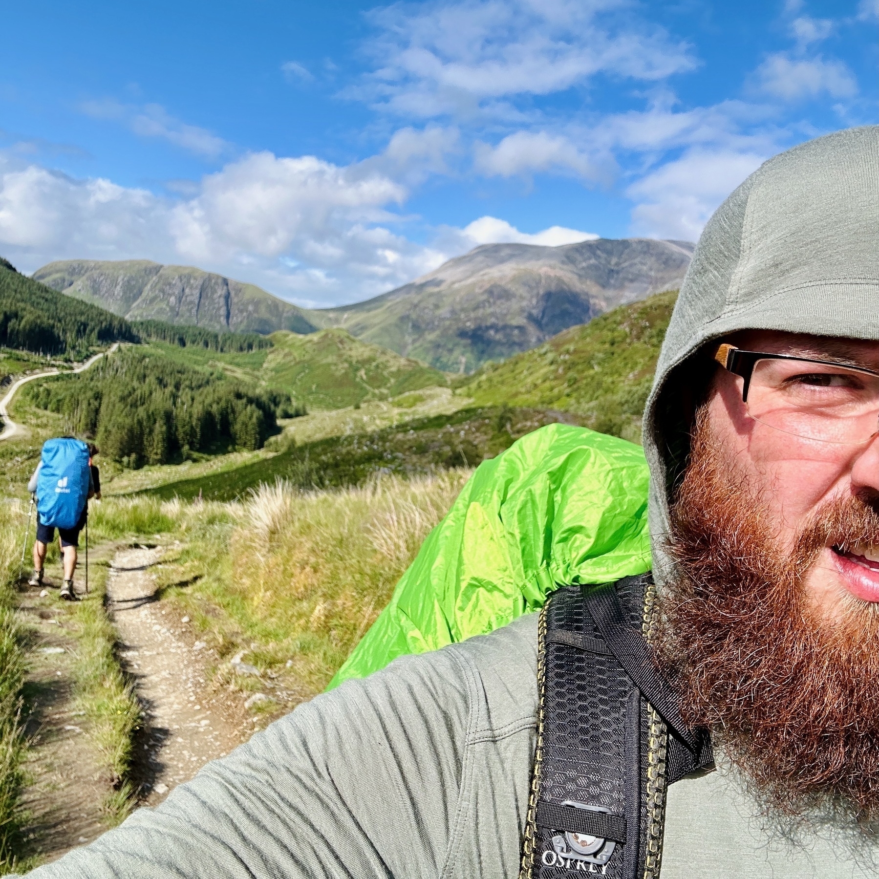 Auto-generated description: Two backpackers are hiking along a mountain trail under a clear blue sky, with one of them looking into the camera.