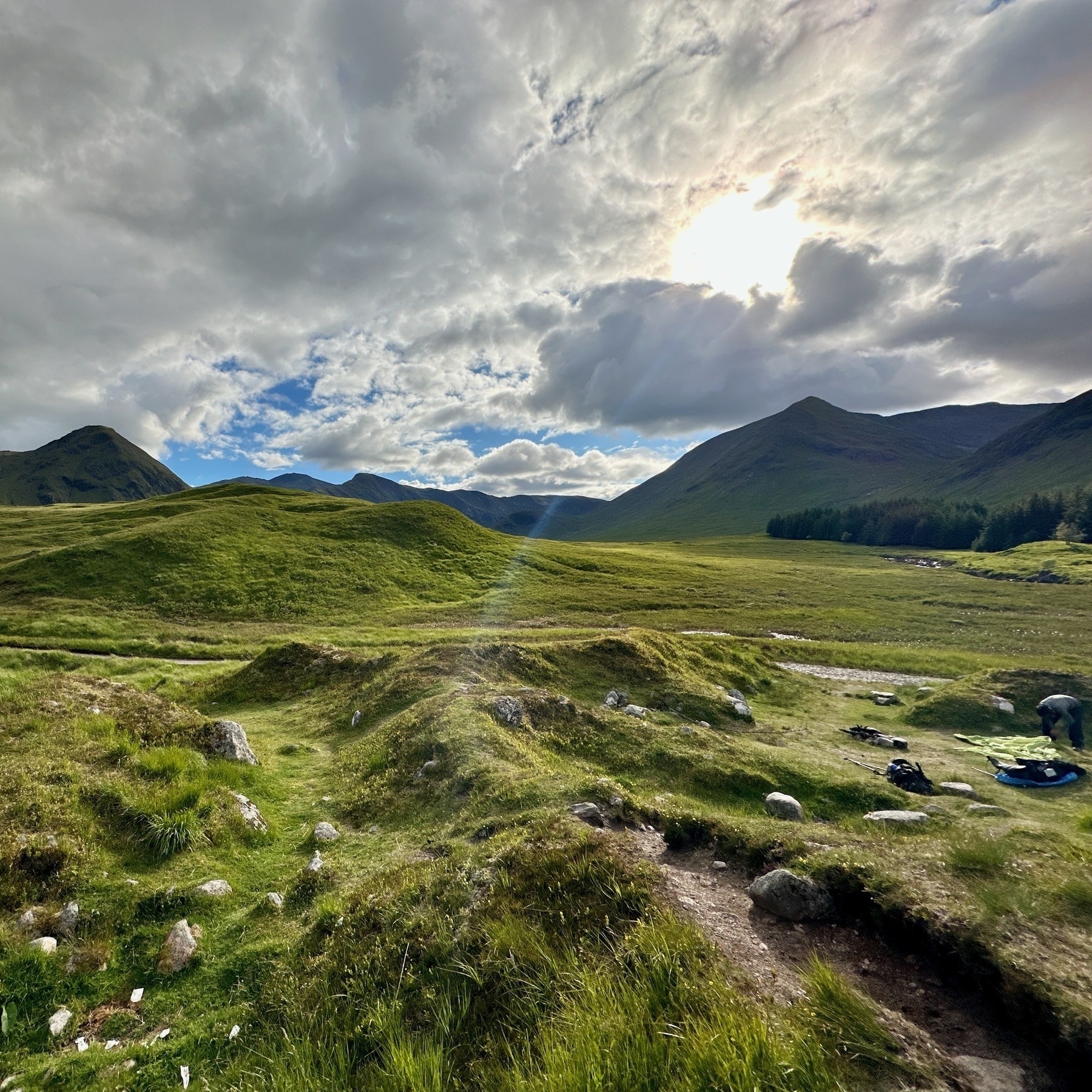 Auto-generated description: A scenic landscape features a grassy valley with rolling hills under a partly cloudy sky, illuminated by the sun.