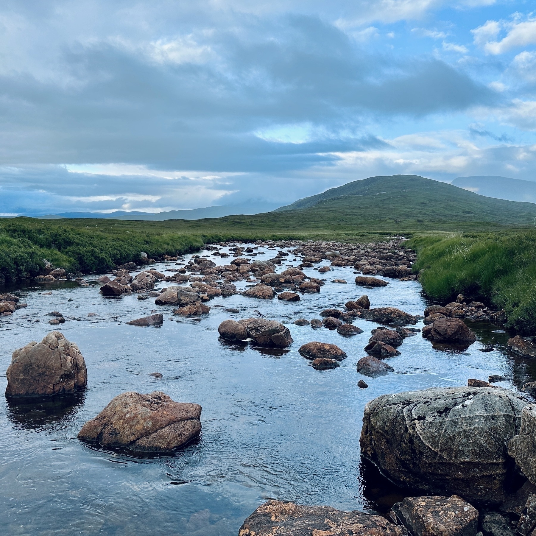 Auto-generated description: A serene landscape features a rocky river flowing through a green field with distant hills under a partly cloudy sky.