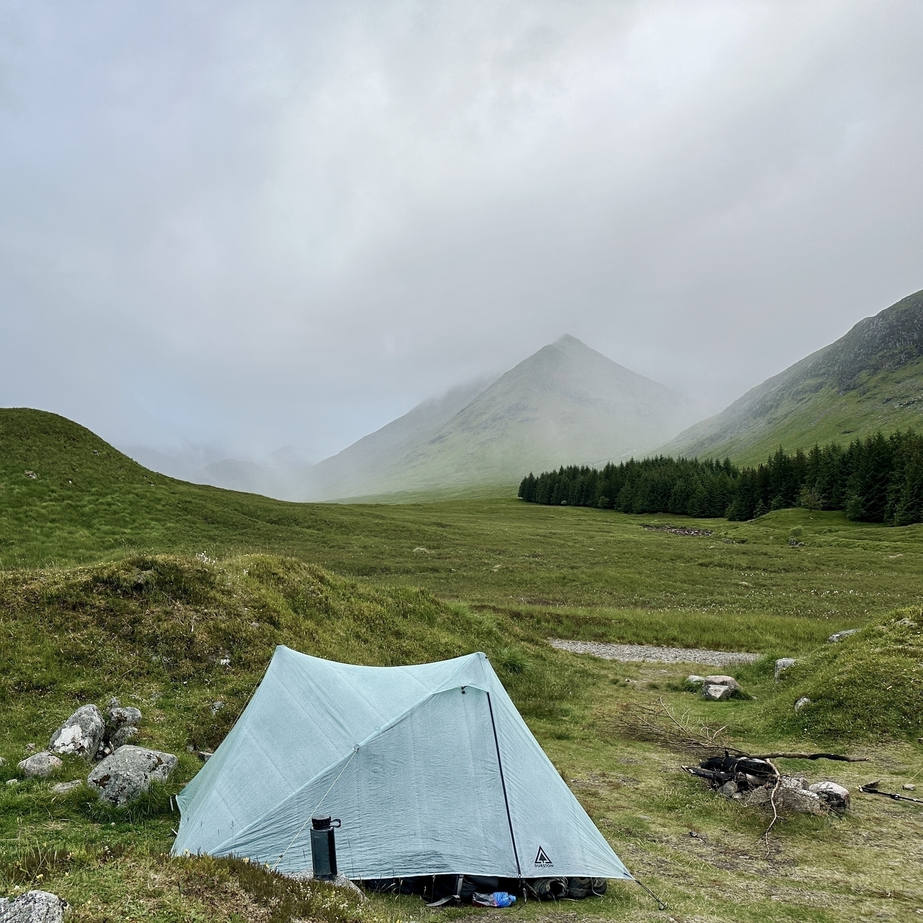 Auto-generated description: A tent is pitched on a grassy field with a misty mountain and a row of trees in the background.