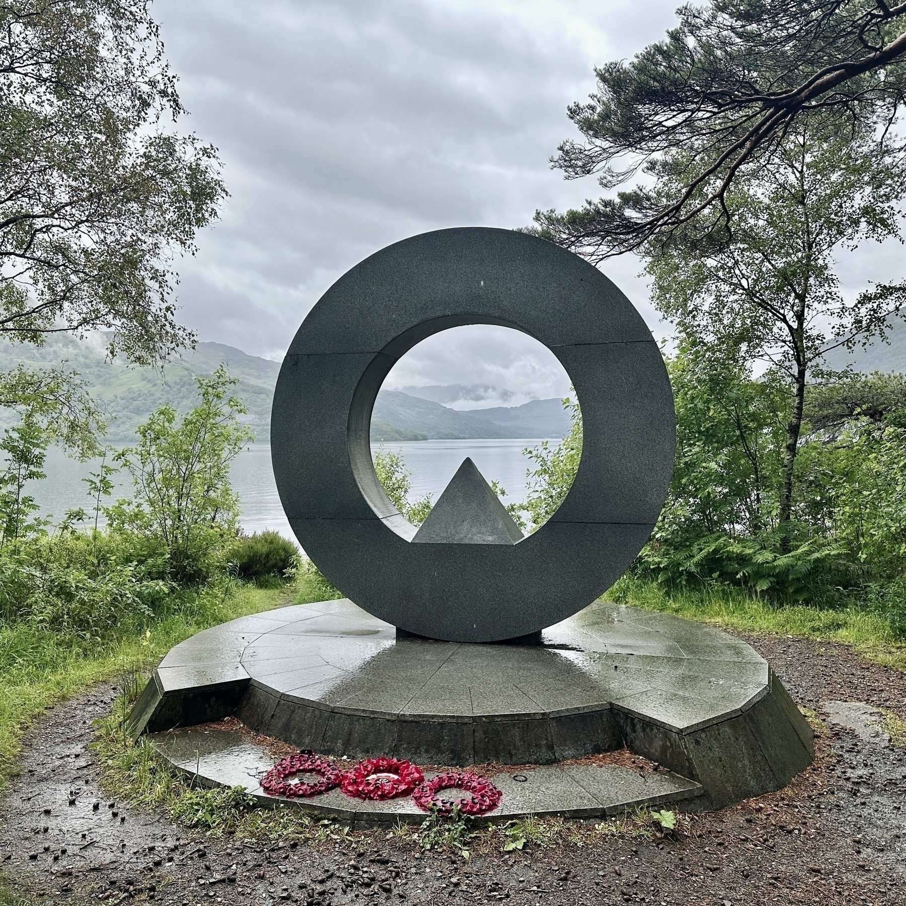 Auto-generated description: A circular monument with a triangular centerpiece stands in a lush, green setting near a body of water, surrounded by trees and featuring flowers placed at its base.