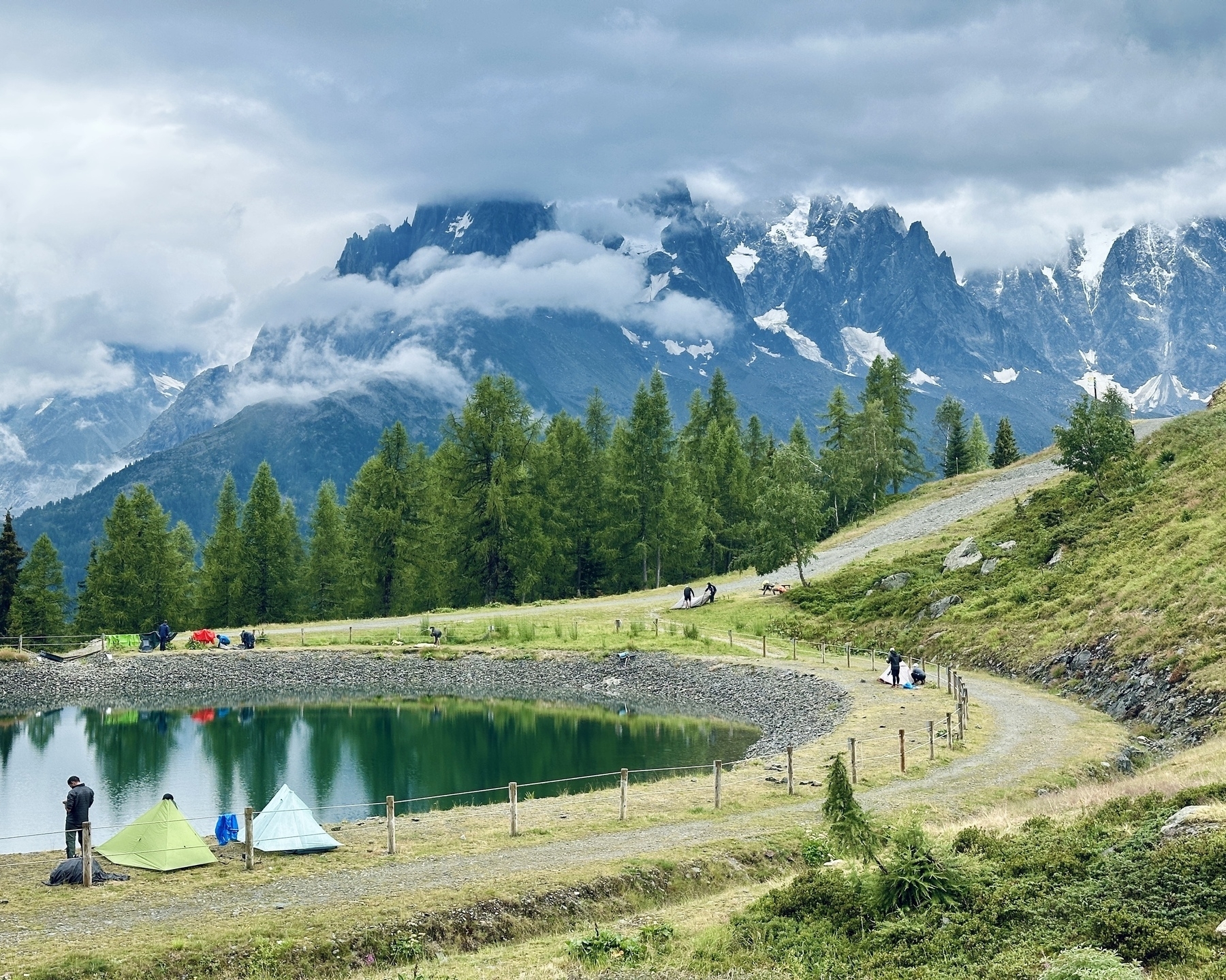 Auto-generated description: A tranquil landscape featuring tents set up by a small pond, surrounded by lush greenery and towering mountains under a cloudy sky.