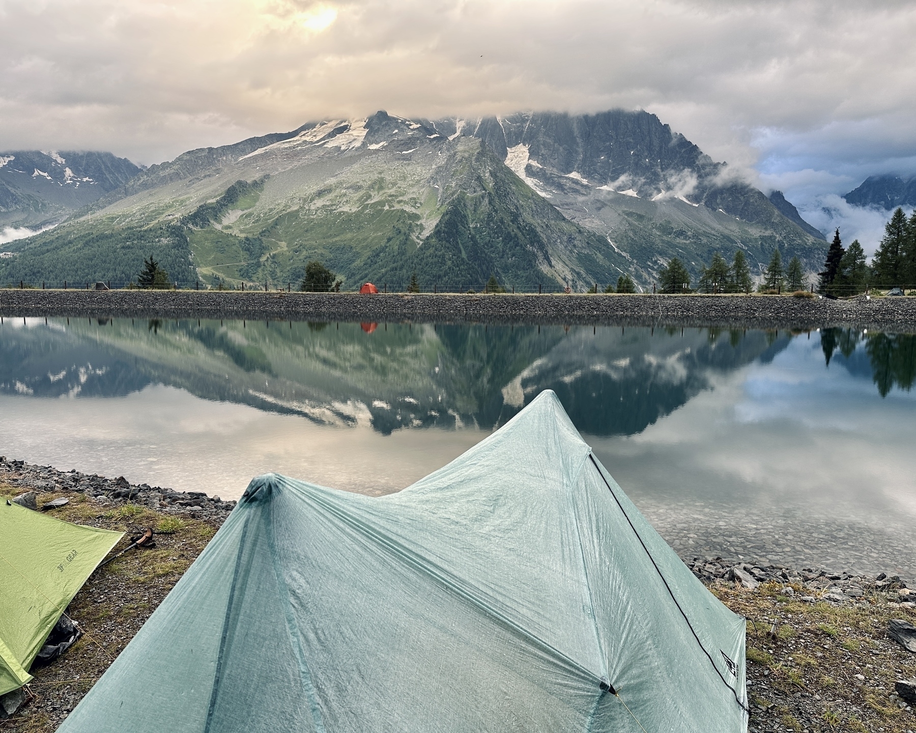 Auto-generated description: Tents are set up beside a tranquil lake reflecting majestic mountains under a cloudy sky.