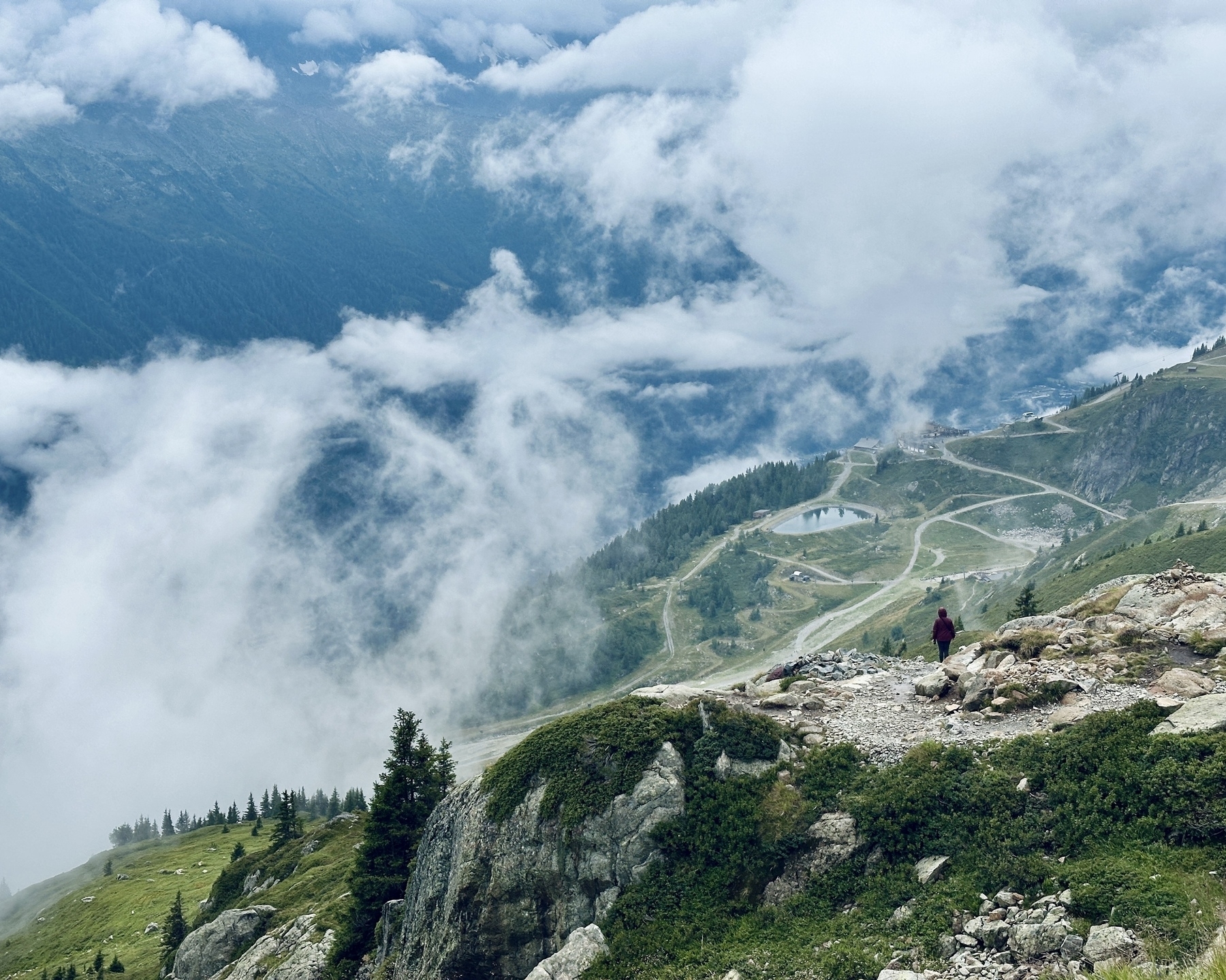 Auto-generated description: A lone hiker stands on a rocky mountain ridge overlooking a valley with winding paths and misty clouds.
