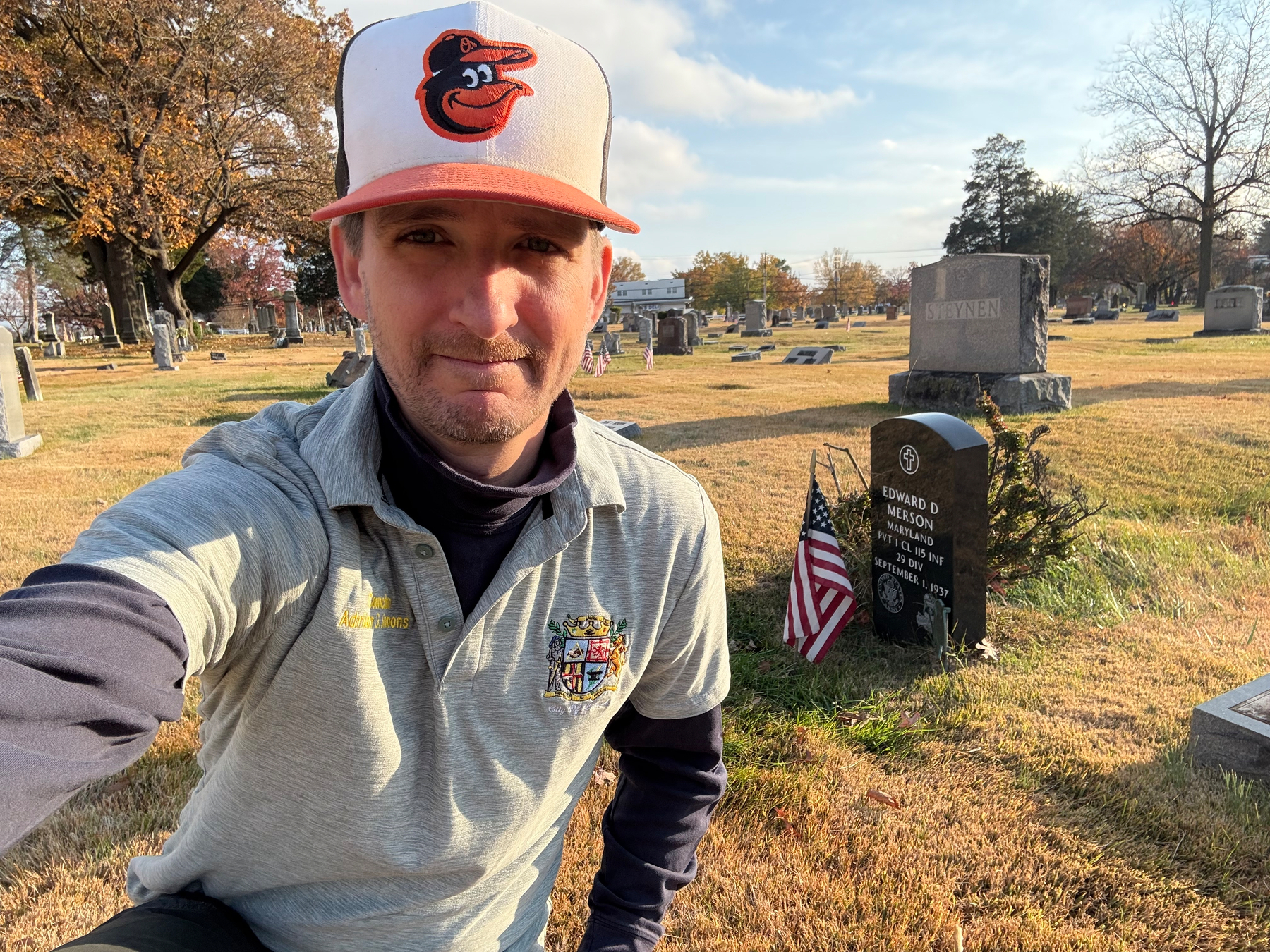 Auto-generated description: A person wearing a baseball cap is taking a selfie in a cemetery next to a gravestone with an American flag placed beside it.