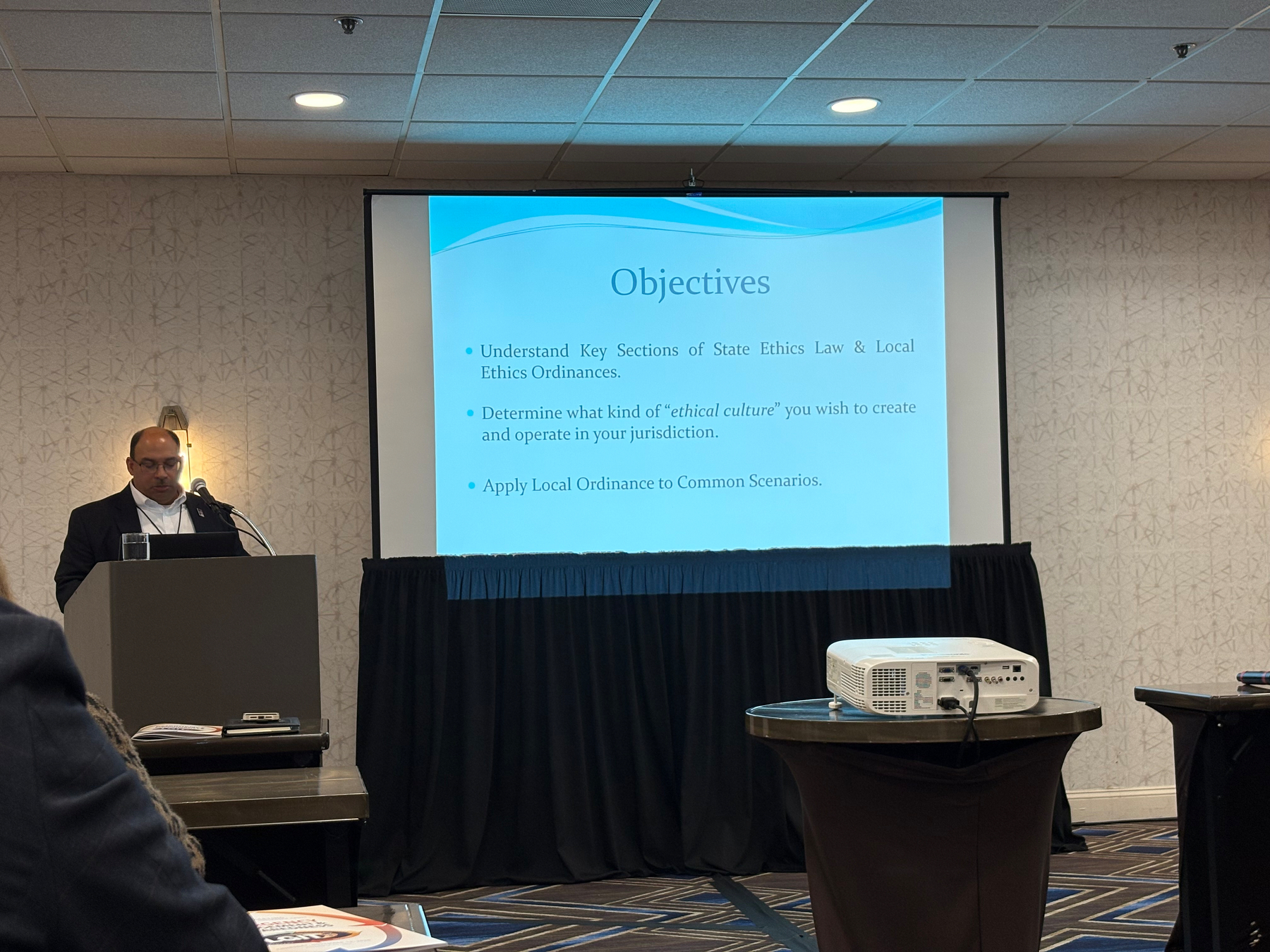 Auto-generated description: A presenter stands at a podium in front of a screen displaying a slide titled Objectives during a seminar.