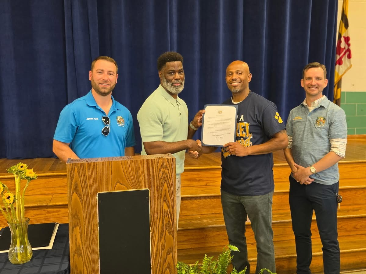 Auto-generated description: Four men are standing on a stage, with two of them shaking hands while holding a certificate.