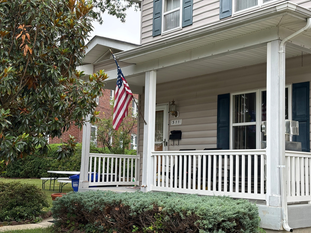 A white house with blue shutters features a porch adorned with an American flag and surrounded by green shrubs and trees.
