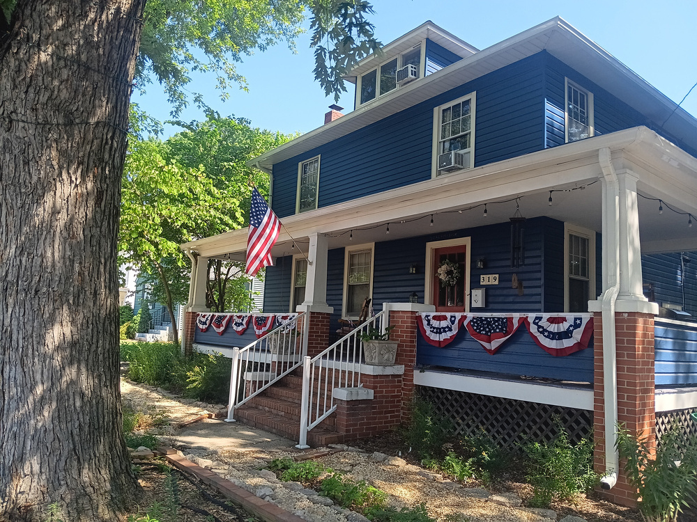 A blue two-story house with a porch decorated with American flags and patriotic bunting is situated next to a large tree.