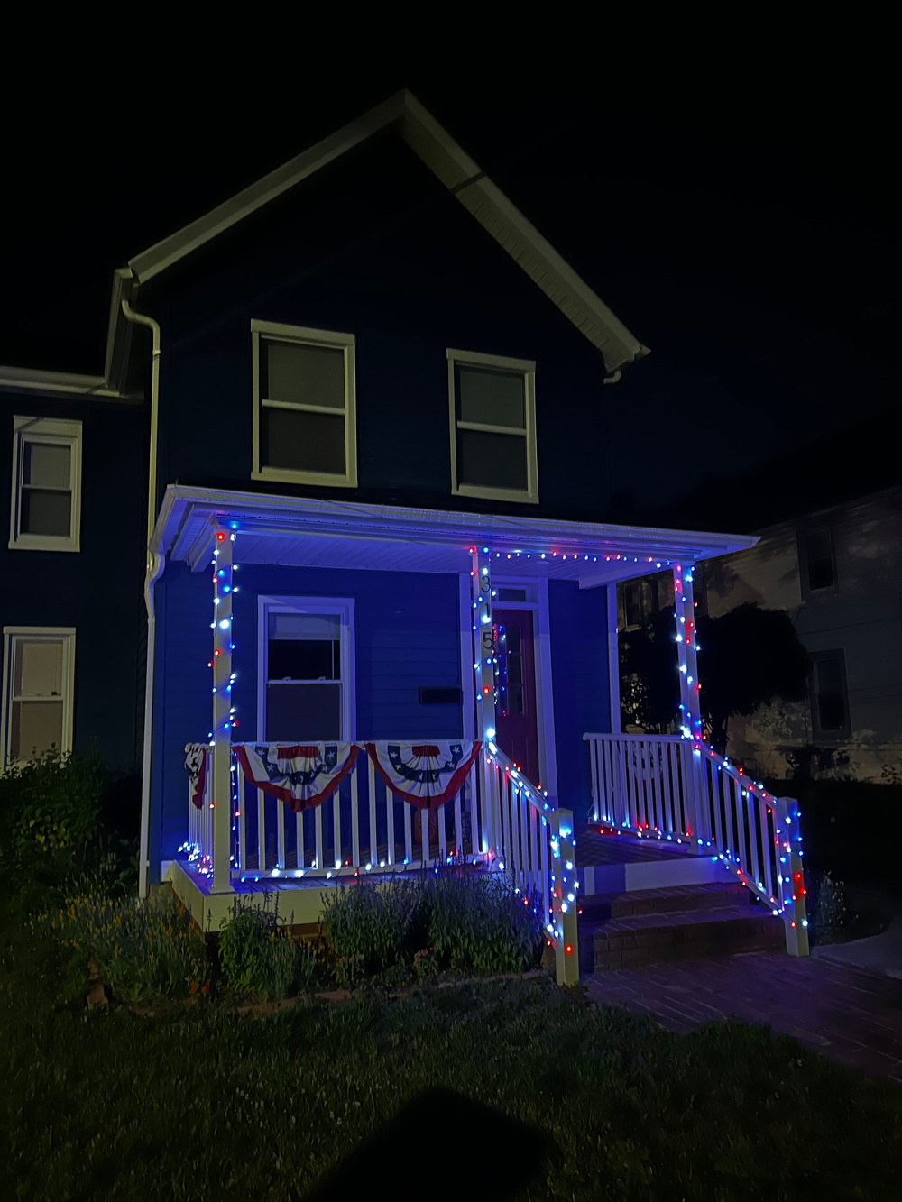 A house porch is decorated with red, white, and blue lights and patriotic bunting against a dark night background.