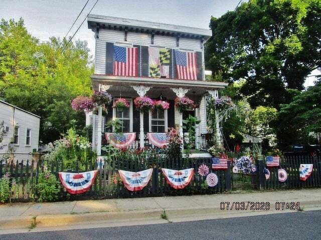 A charming house decorated with American and Maryland flags along with patriotic bunting and flowers is set against a lush green backdrop.