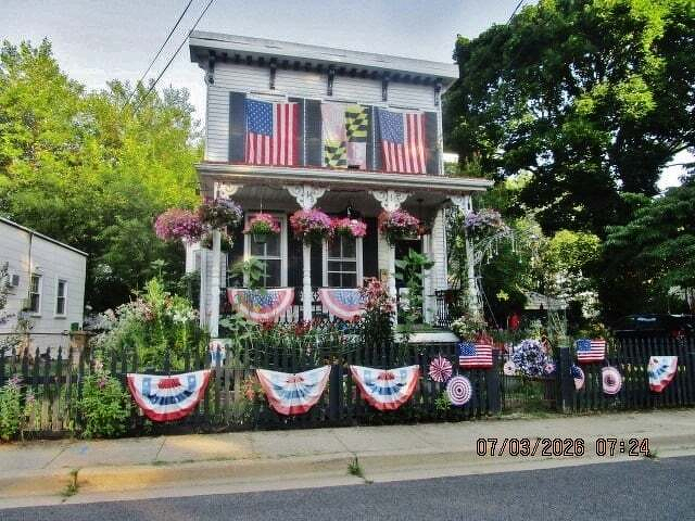 Auto-generated description: A charming house decorated with American and Maryland flags along with patriotic bunting and flowers is set against a lush green backdrop.
