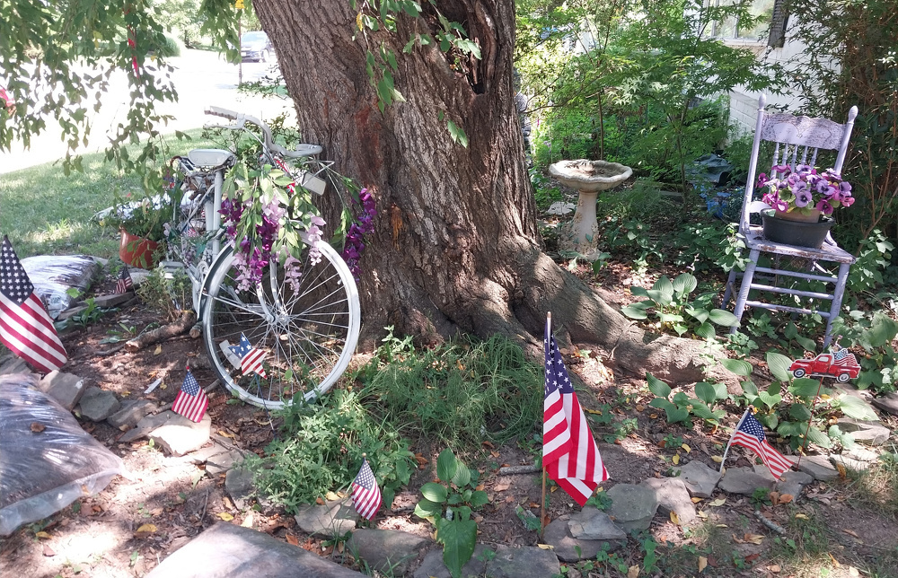 A large tree is surrounded by a decorative arrangement including a white vintage bicycle adorned with purple flowers, several American flags, a birdbath, and a lavender-colored chair holding a planter of flowers.
