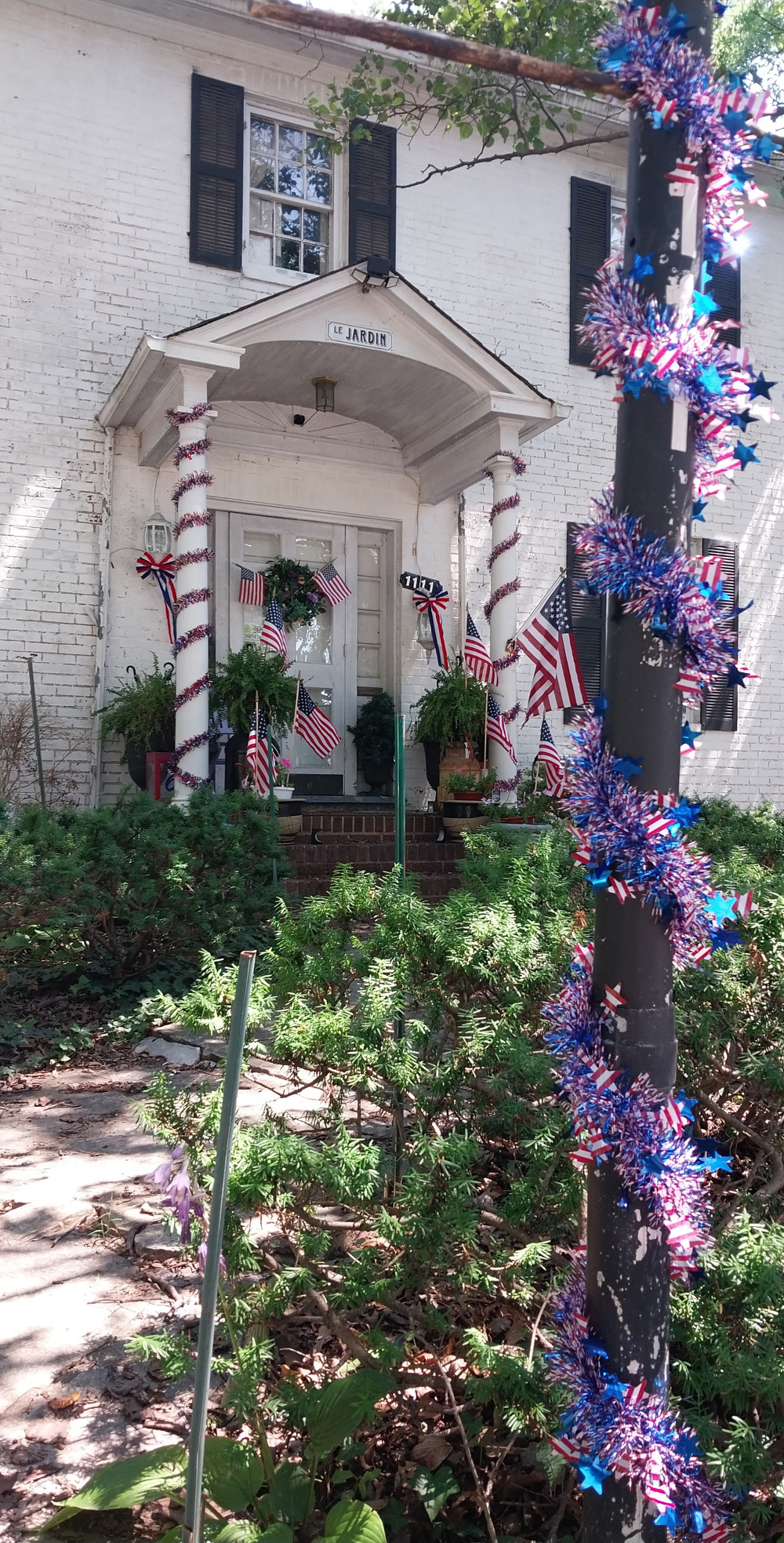 A white brick house entrance is decorated with American flags, red, white, and blue garlands, and greenery, creating a patriotic display.