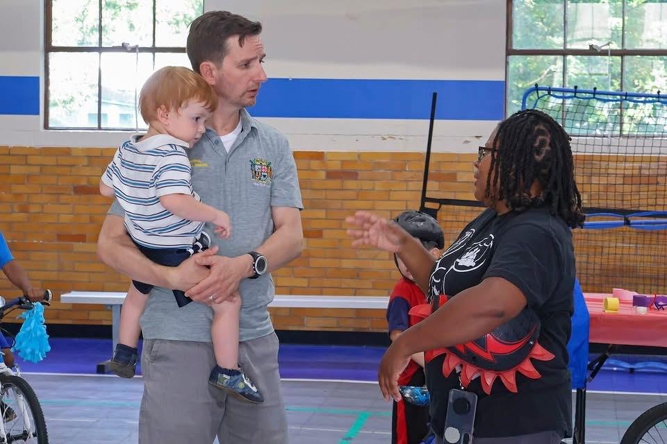Auto-generated description: A man holding a young child converses with a woman in a gymnasium setting where other children and bicycles are also visible.