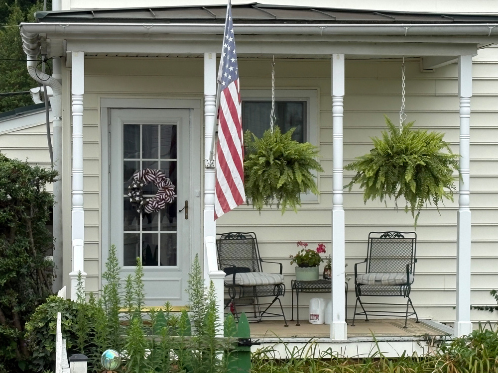 A small porch with two chairs, a table with a potted plant, hanging ferns, and an American flag is attached to a beige house with white trim.