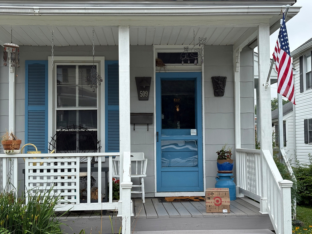 A small front porch with white railings and blue shutters features a blue door, an American flag, and various potted plants and decorations.