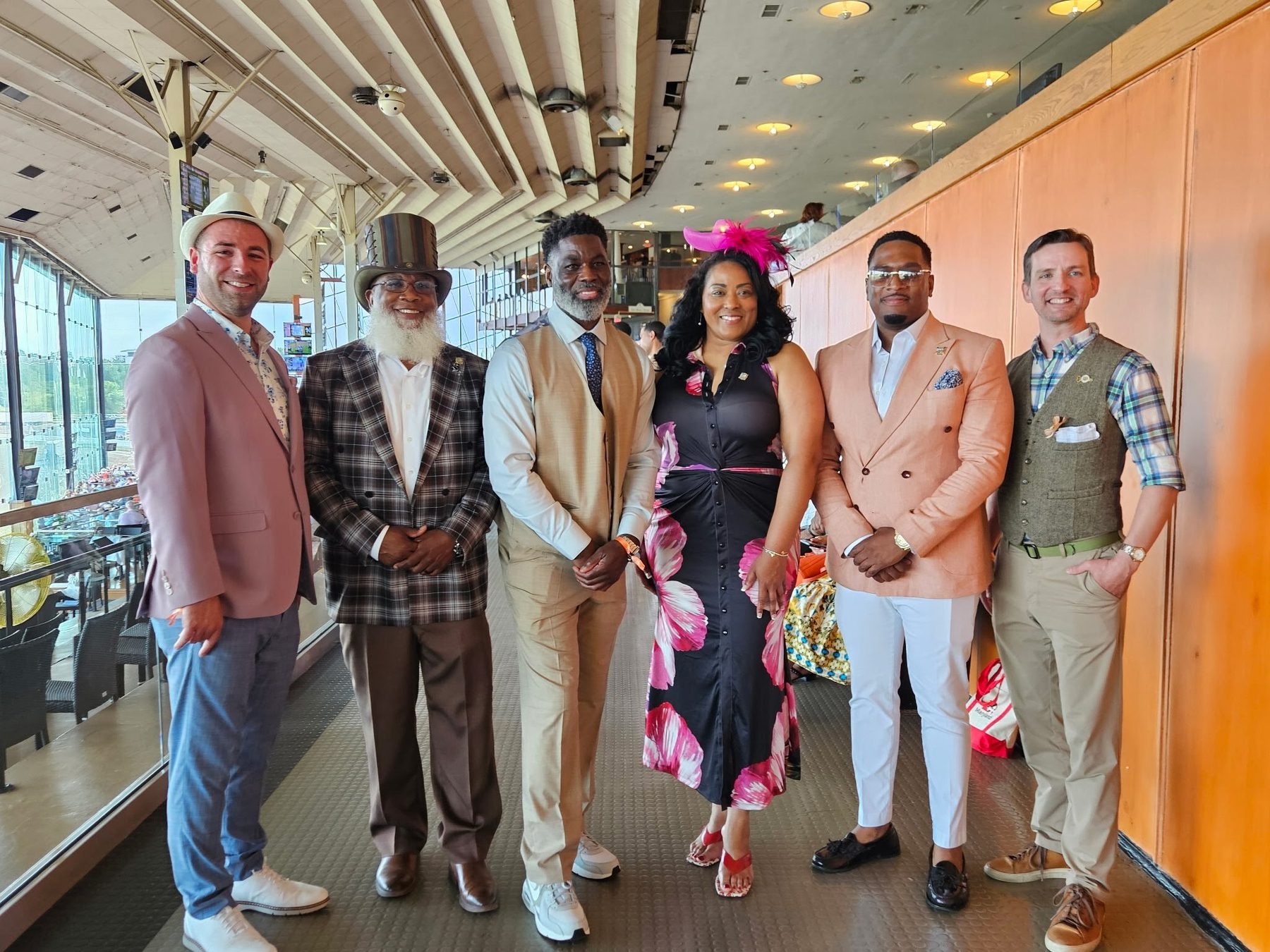 Auto-generated description: A group of six people dressed in stylish, semi-formal attire are standing indoors in a brightly lit space, smiling and posing for the camera.