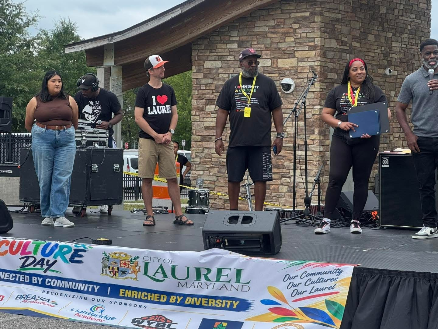 Auto-generated description: A diverse group of five people stand on an outdoor stage during an event celebrating culture and community in Laurel, Maryland.