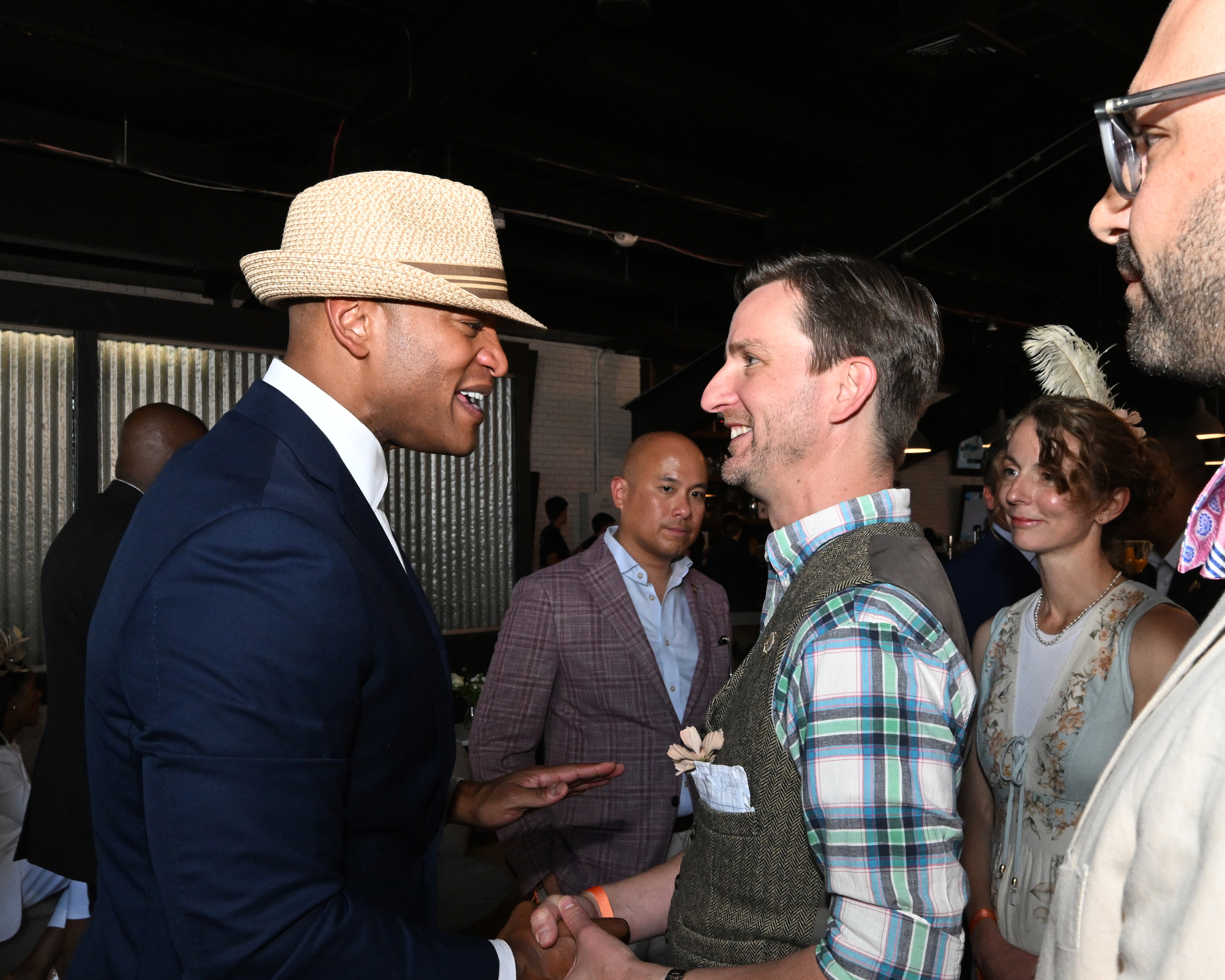 Auto-generated description: A group of people are engaged in friendly conversation and handshakes at an indoor social event.