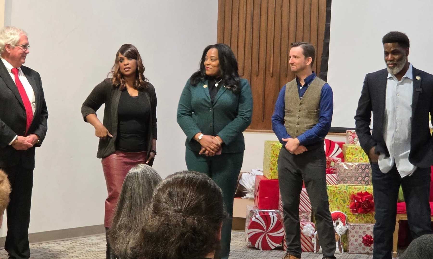 Auto-generated description: Five people are standing in a room, dressed in formal and semi-formal attire, with wrapped gifts in the background.