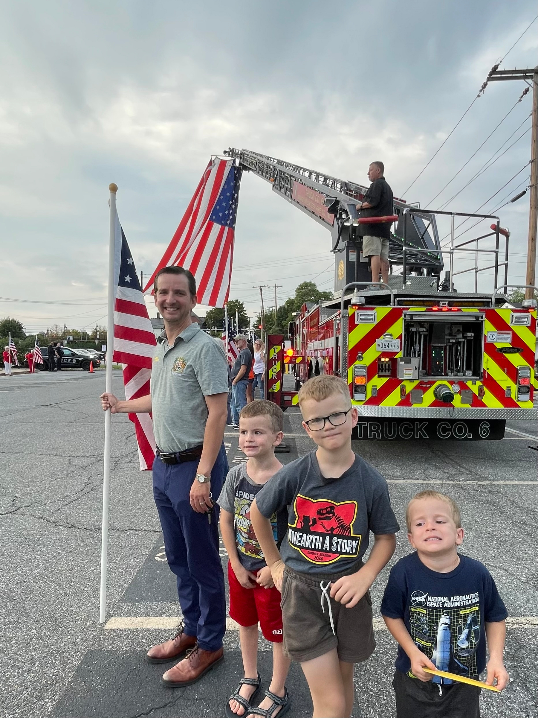 Auto-generated description: A man holding a flag stands with three children in front of a fire truck displaying a large American flag hanging from an extended ladder.