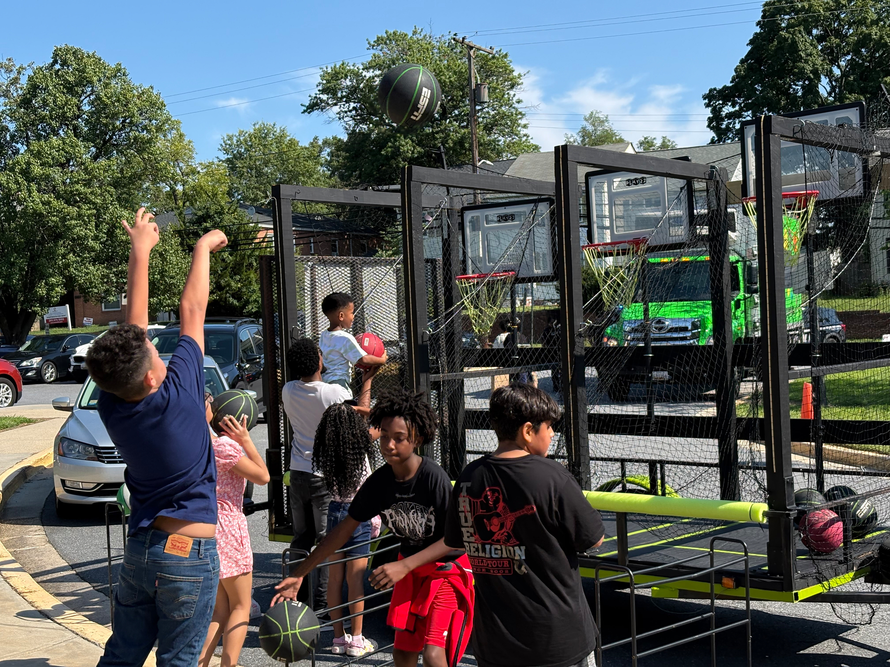 Auto-generated description: A group of children are playing an arcade-style basketball game outdoors on a sunny day.