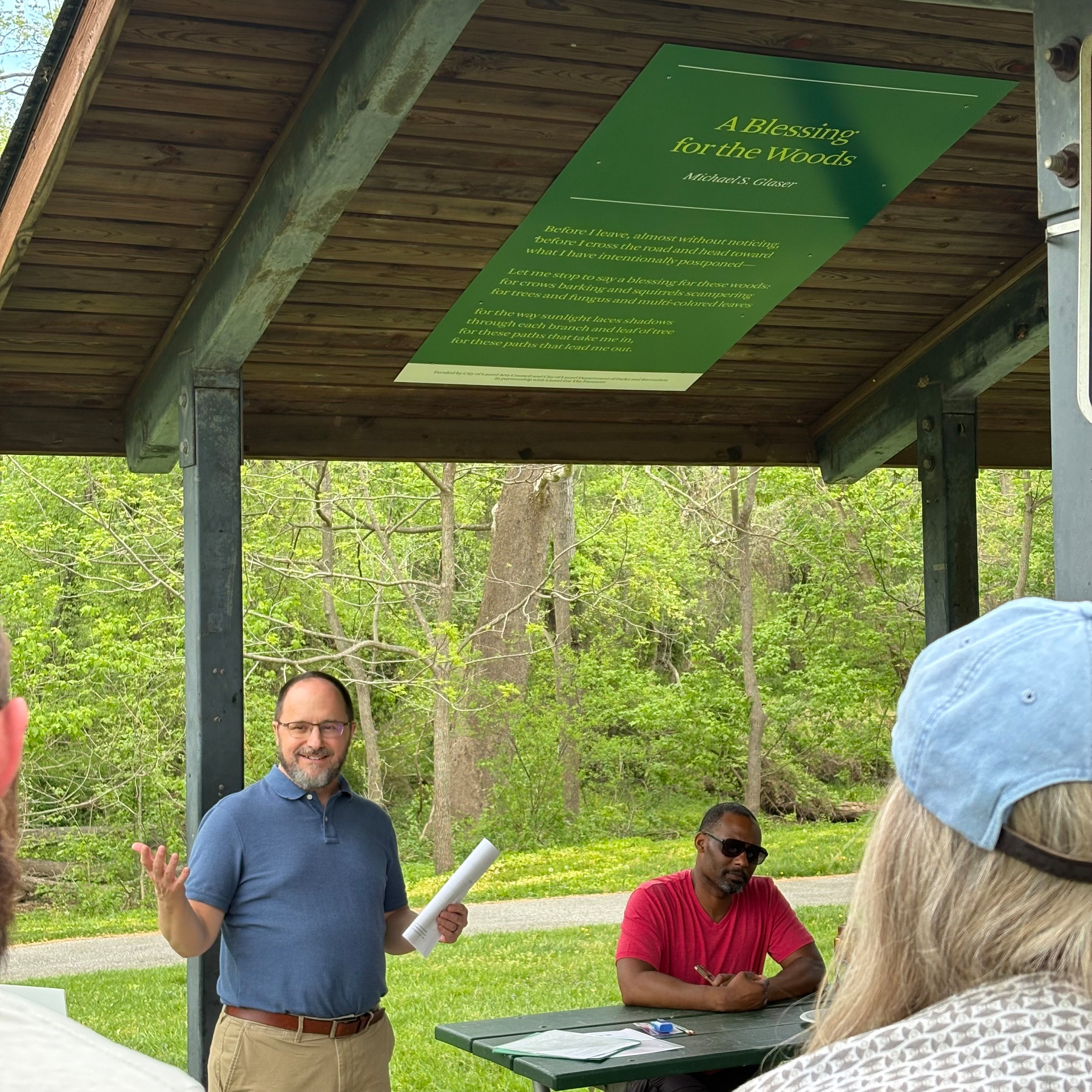 Auto-generated description: A person stands speaking under a covered pavilion while two others sit at a table with lush greenery in the background.