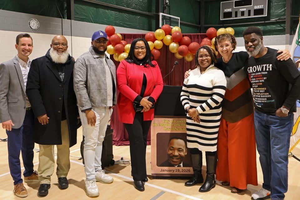 Auto-generated description: A group of seven people is posing together in a gymnasium decorated with a balloon arch and a poster featuring a man's smiling face and the date January 19, 2026.