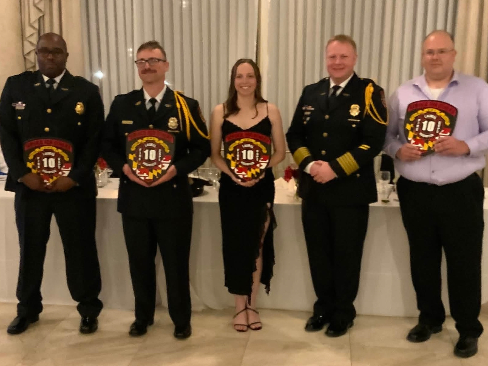 Auto-generated description: Five people, including three in uniform and two in civilian clothing, are standing indoors holding commemorative plaques.