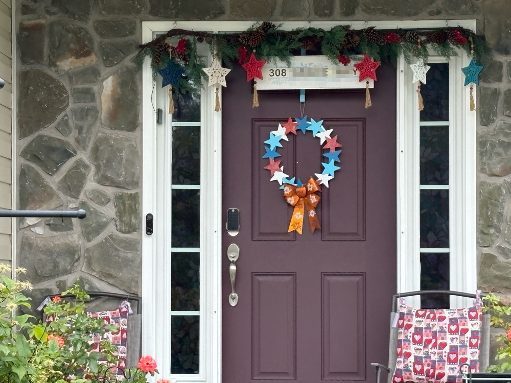 A front door decorated with a colorful star-shaped wreath and a garland of stars and greenery is flanked by two chairs with vibrant patterned cushions.