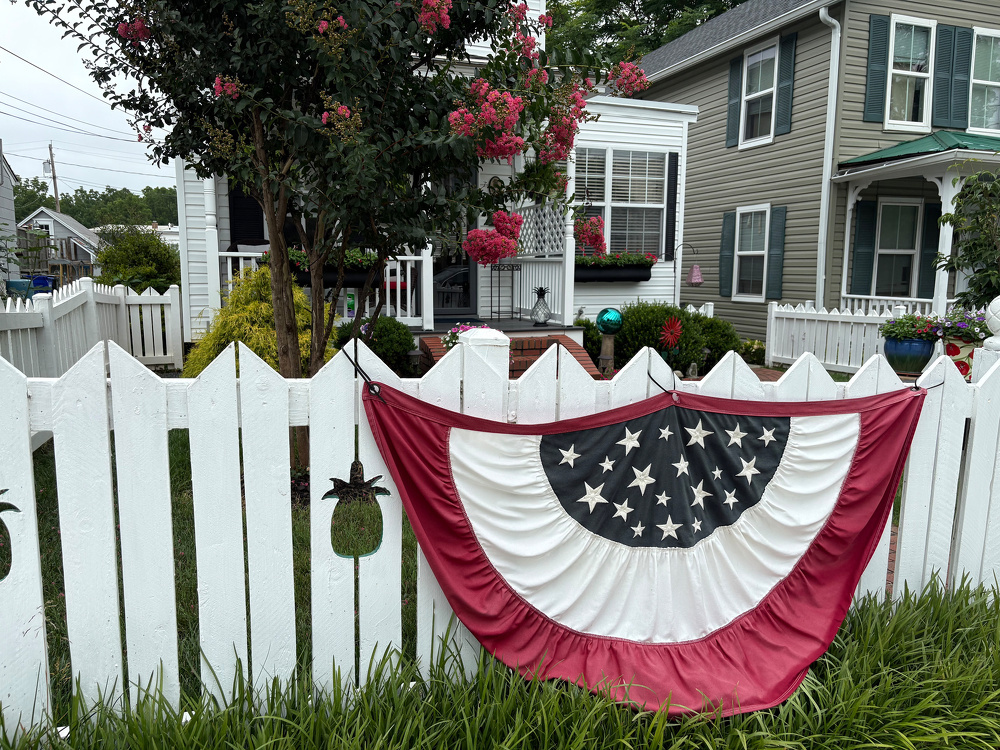A patriotic red, white, and blue bunting is draped over a white picket fence in front of a house with a well-maintained yard and blooming flowers.