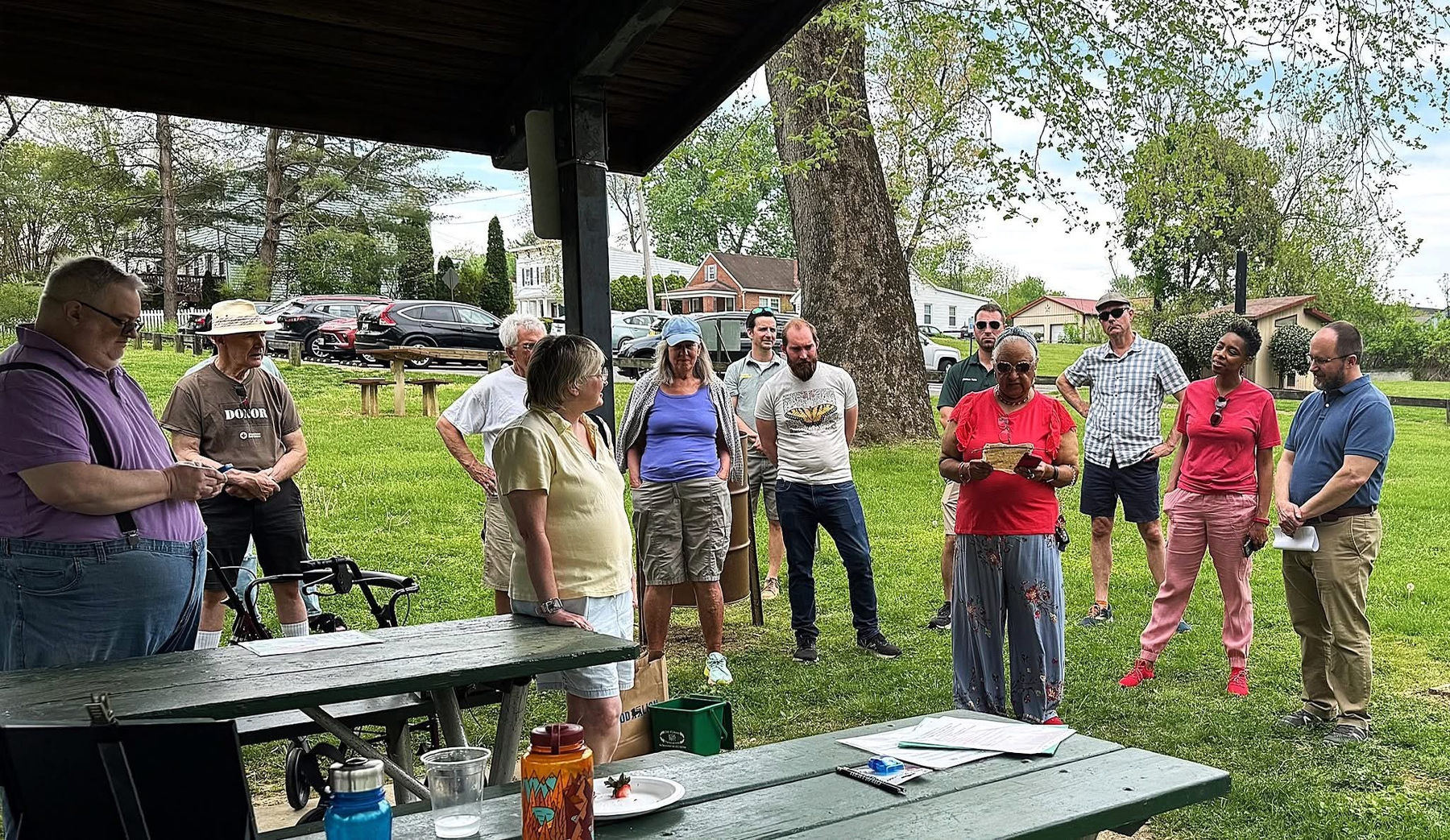 Auto-generated description: A group of people is gathered outdoors under a wooden shelter, some standing and some sitting on picnic benches, engaged in conversation.