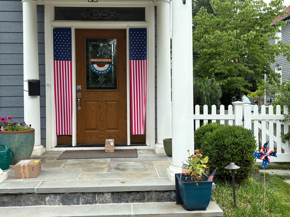 A front porch with a wooden door displaying a Welcome sign, flanked by two American flag decorations, has two delivered packages on the steps and a potted plant with small American flags nearby.