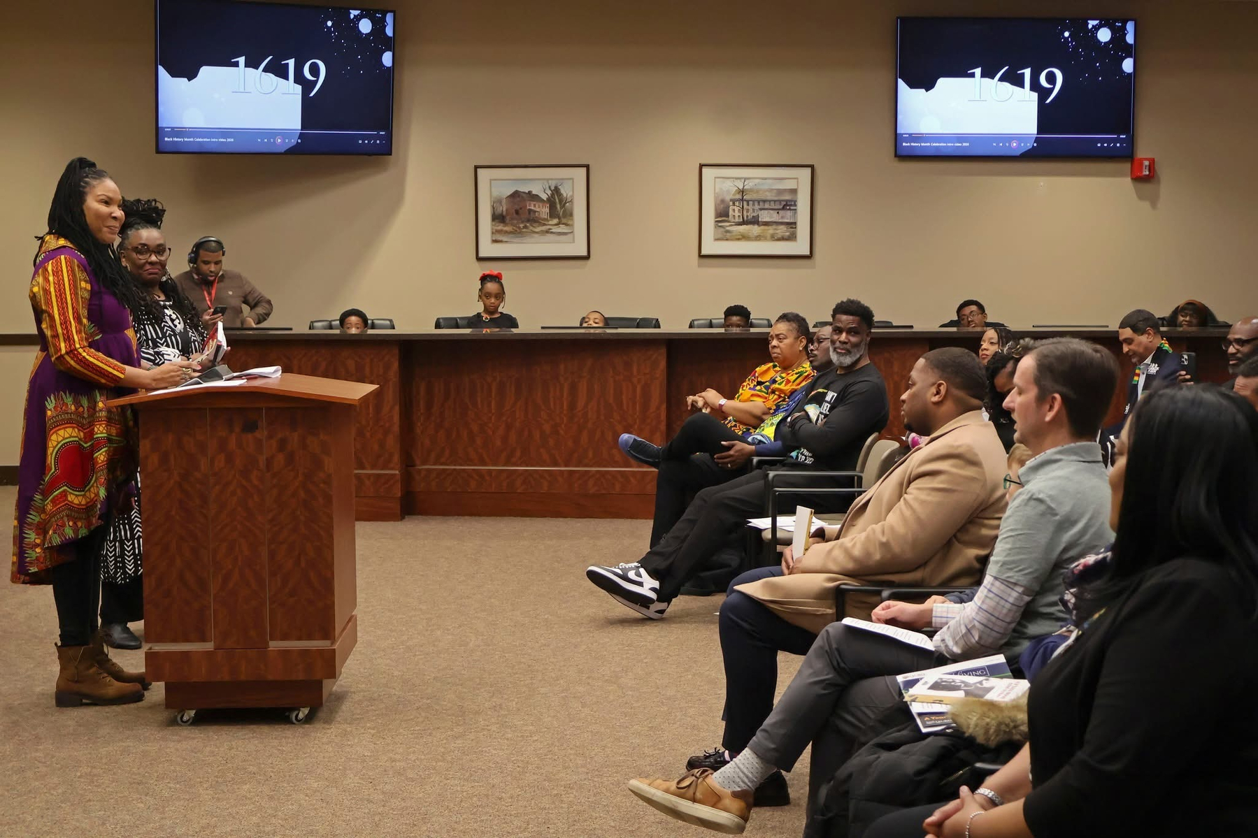 Auto-generated description: A person is speaking at a lectern to an audience in a conference room, with people seated and screens displaying the number 1619.