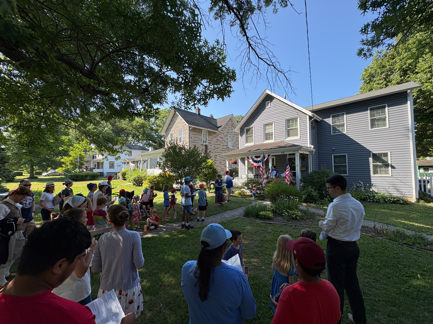 Auto-generated description: A group of people, including adults and children, are gathered outside a house decorated with American flags and bunting, possibly for a community event or celebration.