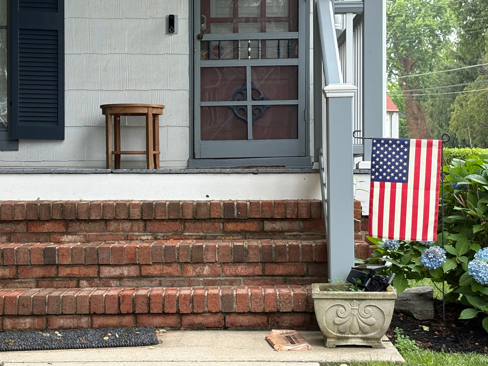 A front porch with brick steps leading up to a door features a small wooden stool, an American flag, and a planter with flowers.